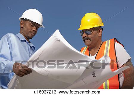Stock Image - Two construction workers discussing blueprints on building site. Fotosearch