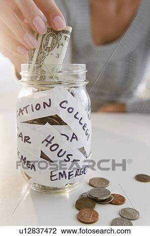 Stock Image - USA, New Jersey, Jersey City, Close-up view of woman's hand putting one dollar bill into piggybank. Fotosearch