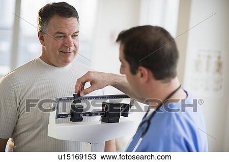 Stock Image - USA, New Jersey, Jersey City, Doctor assisting male patient on weighing scales in hospital. Fotosearch