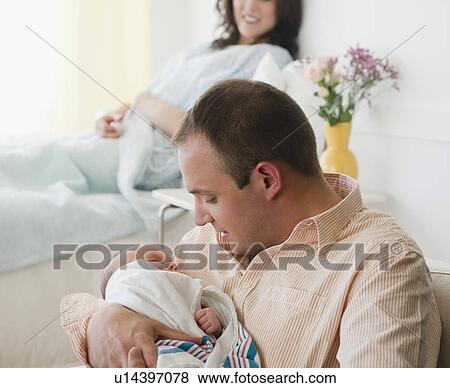 Stock Photo - USA, New Jersey, Jersey City, father embracing baby daughter (2-5 months) with mother in background. Fotosearch