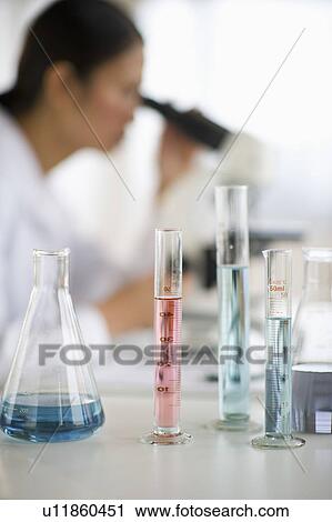 Stock Image - USA, New Jersey, Jersey City, Female scientist using microscope behind test tubes and flasks. Fotosearch