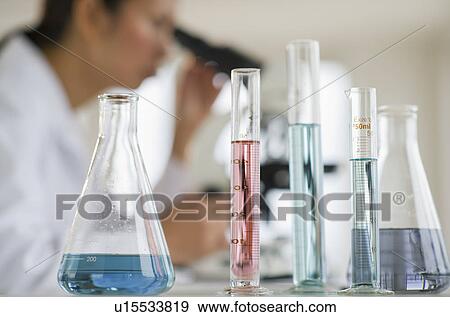 Stock Photo - USA, New Jersey, Jersey City, Female scientist using microscope behind test tubes and flasks. Fotosearch