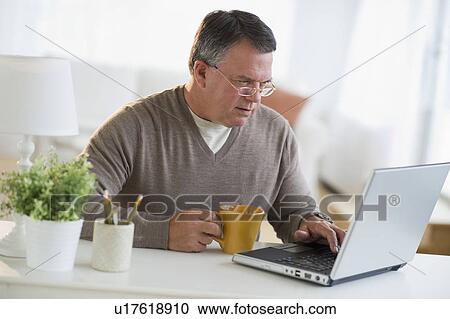Stock Image - USA, New Jersey, Jersey City, Man using laptop, holding drink in living room. Fotosearch