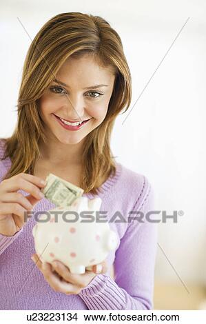 Picture - USA, New Jersey, Jersey City, Portrait of young woman putting banknote to piggybank. Fotosearch