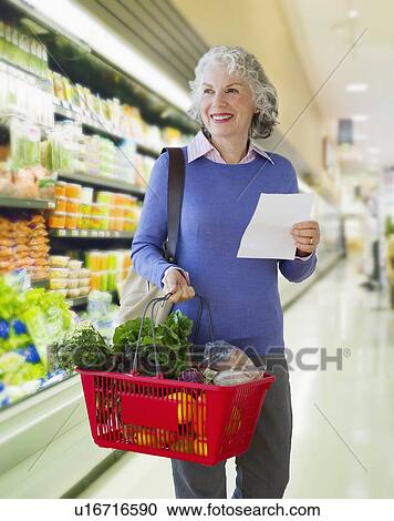 Stock Image - USA, New Jersey, Jersey City, Senior woman carrying shopping basket in supermarket. Fotosearch