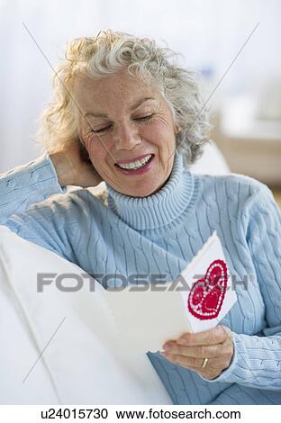 Stock Image - USA, New Jersey, Jersey City, Senior woman reading Valentines card in living room. Fotosearch