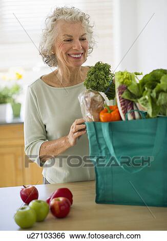 Stock Photograph - USA, New Jersey, Jersey City, Senior woman with grocery bag in kitchen. Fotosearch