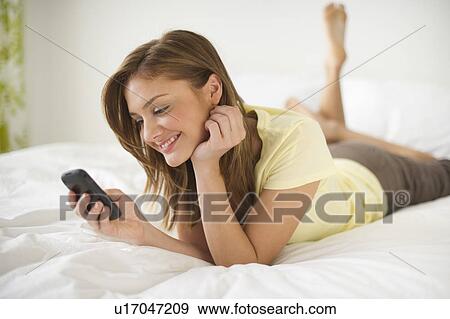 Stock Photo - USA, New Jersey, Jersey City, Young woman lying on bed with mobile phone. Fotosearch