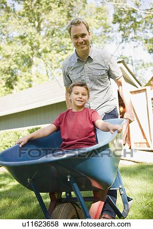 Stock Photo - USA, New York, Flanders, father pushing wheel barrow with son (8-9) inside. Fotosearch