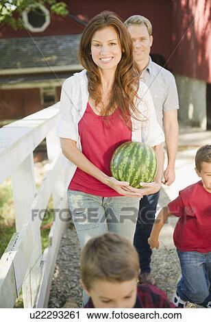 Stock Image - USA, New York, Flanders, Mother holding water melon while boys (4-5, 8-9) are messing about, father in background. Fotosearch
