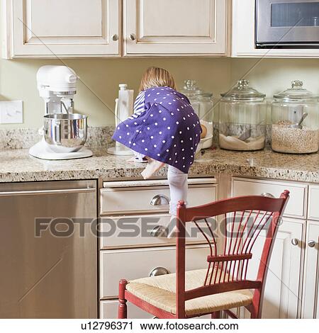 Stock Image - USA, Utah, Girl (2-3) climbing on cupboard in kitchen. Fotosearch