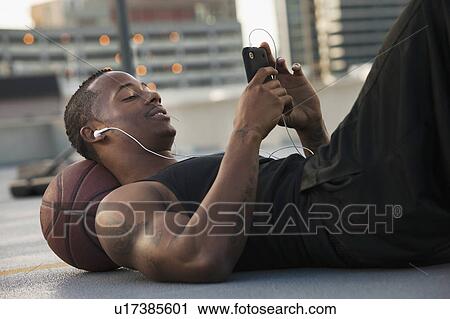 Stock Image - USA, Utah, Salt Lake City, Young man lying on basketball and listening to mp3 player. Fotosearch