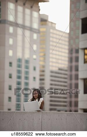 Stock Image - USA, Utah, Salt Lake City, Young businesswoman using laptop in front of skyscrapers. Fotosearch