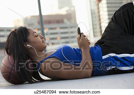 Picture - USA, Utah, Salt Lake City, Young woman lying on basketball and listening to mp3 player. Fotosearch