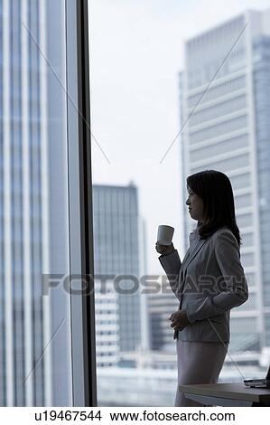 Businesswoman standing by window and holding a cup of coffee View Large Photo Image Picture - Businesswoman standing by window and holding a cup of coffee. Fotosearch