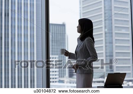 Stock Image - Businesswoman standing by window and holding a cup of coffee. Fotosearch