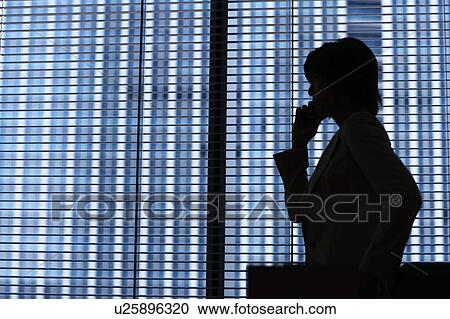 Stock Image - Silhouette of businesswoman standing by window and on cell phone. Fotosearch