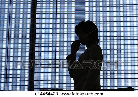 Stock Photo - Silhouette of businesswoman standing by window and on cell phone. Fotosearch