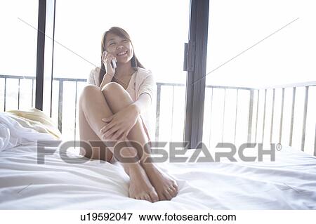 Stock Photo - Young woman sitting on bed using cell phone. Fotosearch