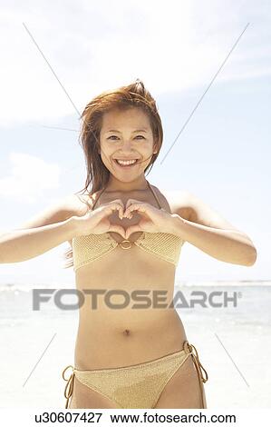Stock Photo - Young woman standing on beach making heart with hands, Guam, USA. Fotosearch