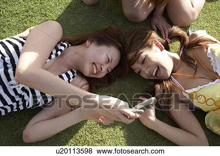 Stock Photo - Young women lying on grass looking at cell phone, Guam, USA. Fotosearch
