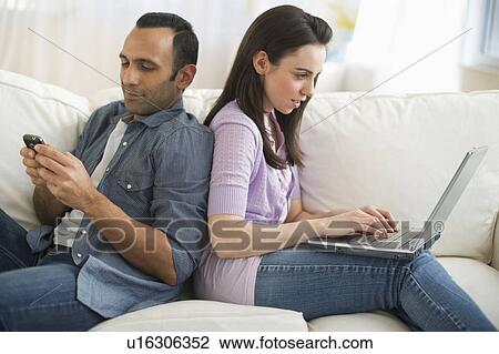 Stock Image - Couple sitting on sofa back to back using laptop and phone. Fotosearch