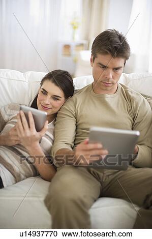 Stock Image - Couple sitting on sofa, using digital tablets. Fotosearch