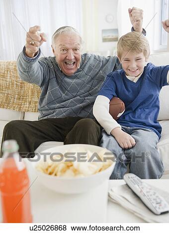 Stock Photo - Grandfather and grandson (8-9) watching tv and cheering. Fotosearch