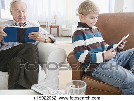 Stock Image - Grandfather reading book, grandson (8-9) reading e-book in living room. Fotosearch