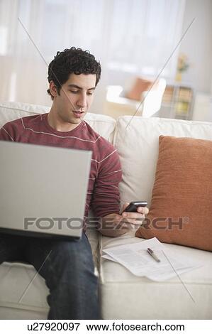 Stock Photo - Man dialing and using laptop on sofa. Fotosearch