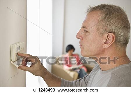 Stock Image - Mature man adjusting room temperature, while woman is sitting in background. Fotosearch