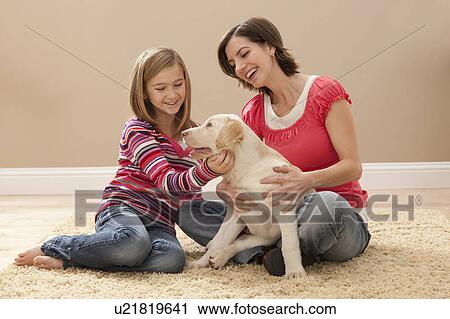 Stock Image - Mother and daughter (10-11) playing with Labrador on carpet. Fotosearch