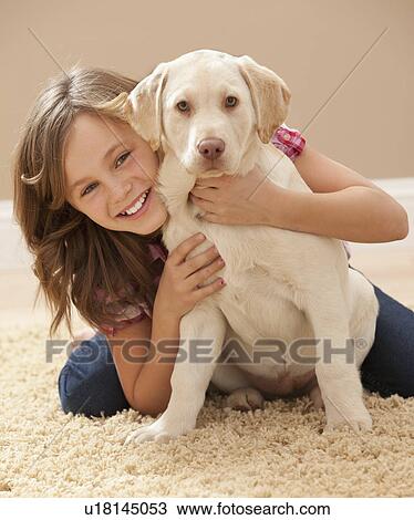 Stock Image - Portrait of girl (10-11) embracing Labrador on carpet. Fotosearch