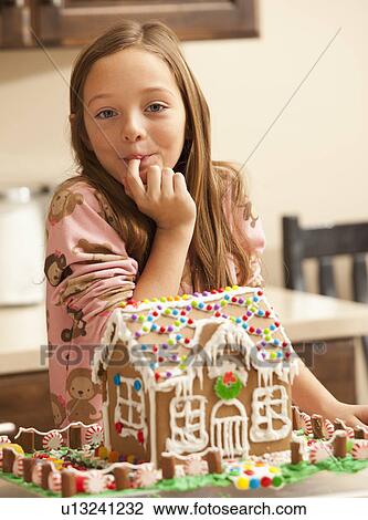 Stock Image - Portrait of girl (6-7) licking finger by gingerbread house. Fotosearch