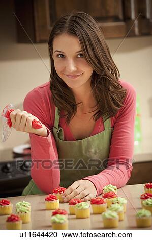 Stock Image - Portrait of young woman decorating cupcakes in kitchen. Fotosearch
