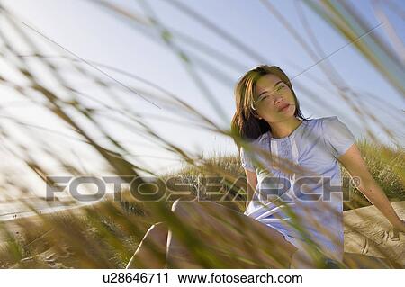 Stock Image - USA, California, Point Reyes, Young woman sitting in grass on sand dune. Fotosearch