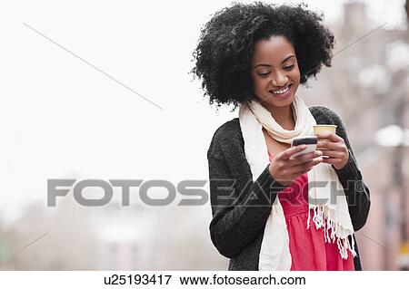 Stock Photo - USA, Washington State, Seattle, Cheerful young woman text messaging while holding takeaway coffee. Fotosearch