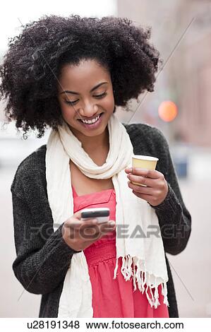 Stock Photo - USA, Washington State, Seattle, Cheerful young woman text messaging while holding takeaway coffee. Fotosearch
