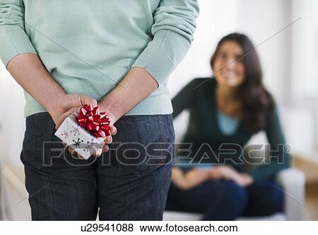 Stock Photo - Young man with present behind his back, girlfriend in background. Fotosearch