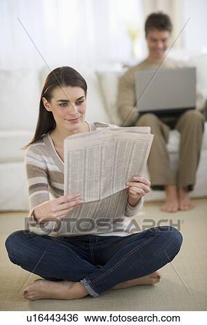 Stock Photograph - Young woman reading newspaper while man is using laptop in background. Fotosearch