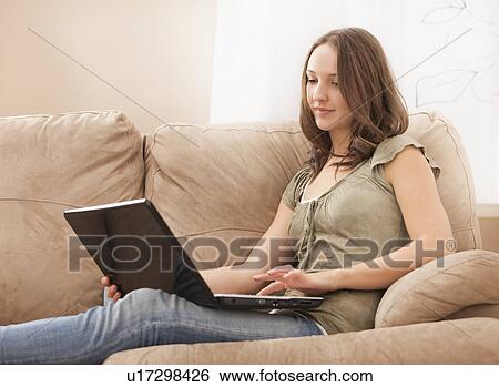 Stock Photograph - Young woman using laptop while sitting at sofa. Fotosearch
