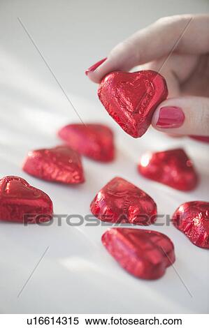 Stock Photography - Close up of woman's fingers with red nail polish holding chocolate in shape of heart in red wrapping. Fotosearch