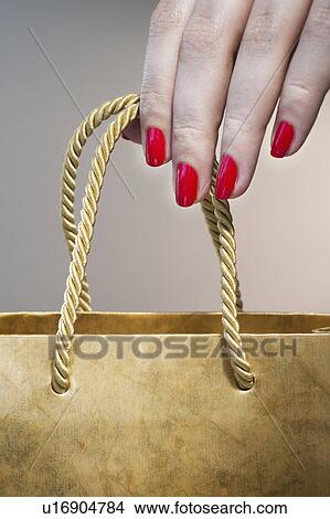 Picture - Close up of woman's hand with red nail polish holding string handle of paper bag. Fotosearch