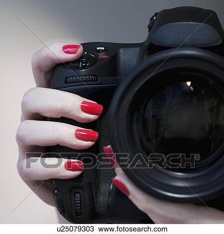 Stock Image - Close up of woman's hand with red nail polish holding camera. Fotosearch