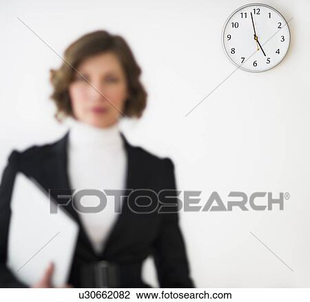 Stock Image - Defocused businesswoman with office clock showing time in background. Fotosearch