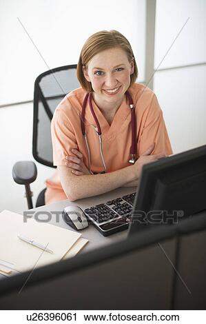 Stock Image - Female nurse sitting at desk and smiling. Fotosearch