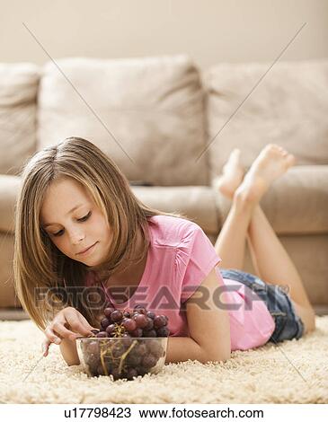 Stock Image - Girl (10-11) lying on floor eating grapes. Fotosearch