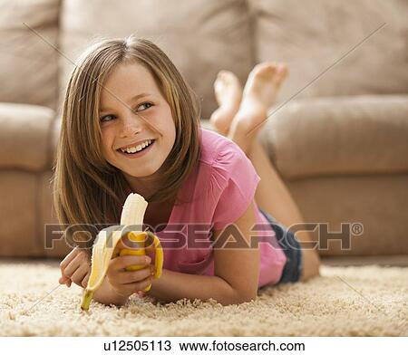 Stock Image - Girl (10-11) lying on rug, eating banana. Fotosearch