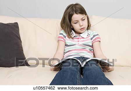 Stock Image - Girl (6-7) reading book on sofa. Fotosearch