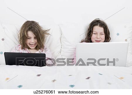 Stock Image - Girls (6-7, 8-9) with laptops sitting in bed. Fotosearch